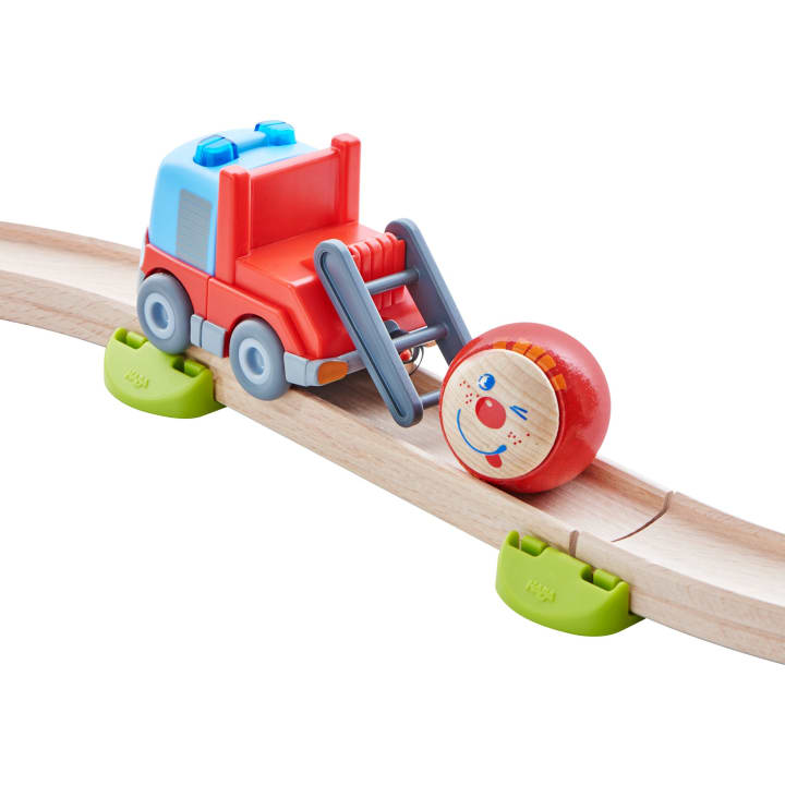 Ein roter und blauer Spielzeug-LKW mit einem grauen Frontlader fährt auf einer Holzeisenbahnstrecke und schiebt einen runden Holzklotz mit einem aufgemalten lächelnden Gesicht. Das Gleis wird von zwei grünen Blöcken gestützt.