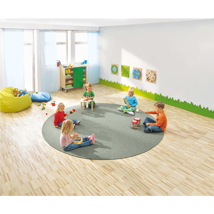 Fünf kleine Kinder sitzen auf einem runden grauen Teppich in einem hellen Klassenzimmer und spielen mit Musikinstrumenten. Im Hintergrund stehen Spielzeuge und ein Sitzsack, und an einer weißen Wand mit grüner Umrandung hängen bunte Wanddekorationen.