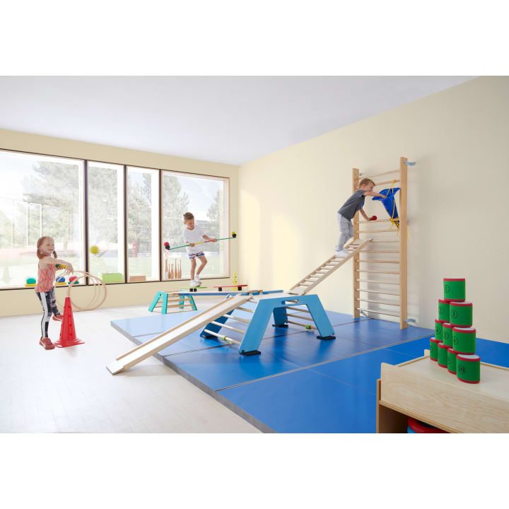 Drei Kinder spielen in einer hellen Turnhalle mit weichen blauen Matten, Schwebebalken und Klettergeräten. Ein Kind klettert auf eine Leiter, ein anderes balanciert auf einem Balken, und ein drittes benutzt Ringe. Ein Stapel grüner und roter Becher steht auf einem Tisch.