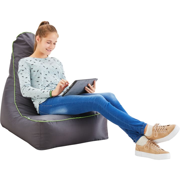 Eine junge Frau sitzt auf einem grauen Sitzsack, trägt einen hellblau gemusterten Pullover, blaue Jeans und hellbraune Turnschuhe, während sie ein Tablet benutzt und lächelt.
