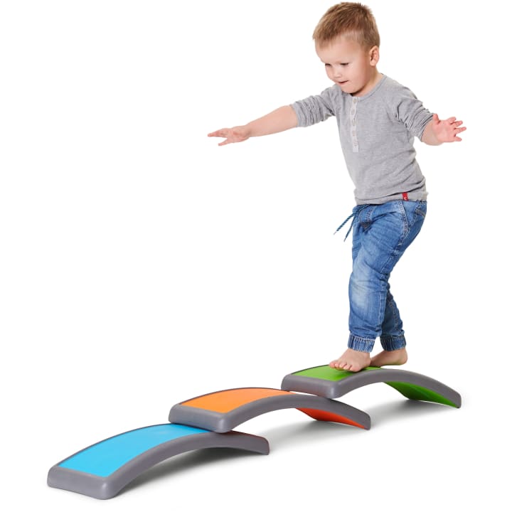 Ein kleines Kind mit blondem Haar, das ein graues Hemd und eine blaue Jeans trägt, balanciert barfuß auf geschwungenen, bunten Trittsteinen, die in einer Reihe auf einem weißen Hintergrund angeordnet sind.