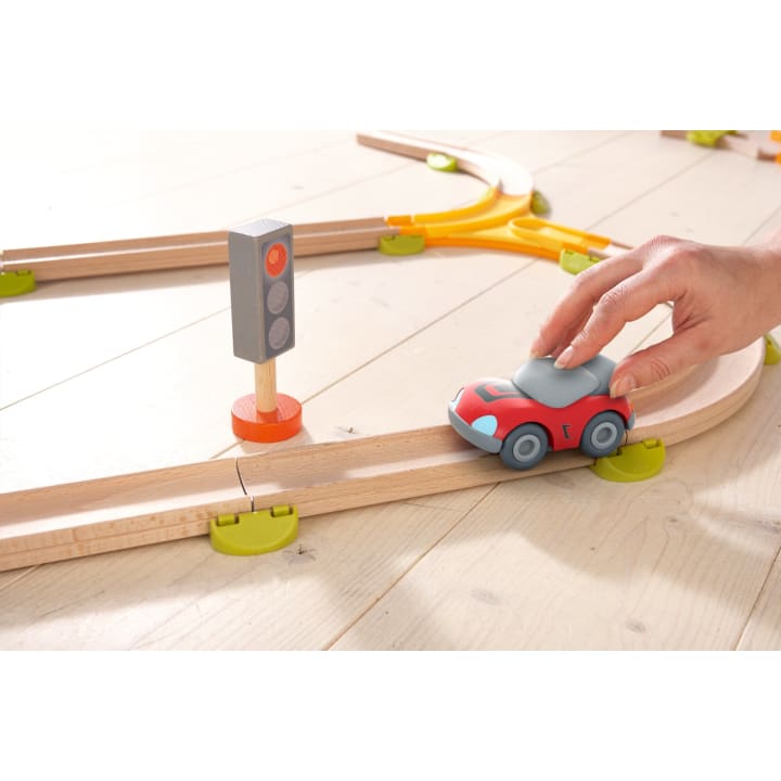 Eine Hand schiebt ein kleines rotes Spielzeugauto auf einer Holzschiene neben einer Spielzeugampel mit roten, gelben und grünen Lichtern auf einer hellen Holzfläche.