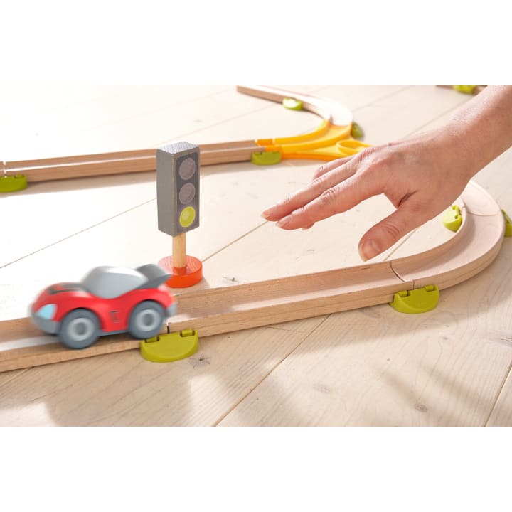 Eine Hand greift nach einem roten Spielzeugauto auf einer Holzeisenbahn, daneben steht eine kleine Spielzeugampel, alles auf einem hellen Holzboden.
