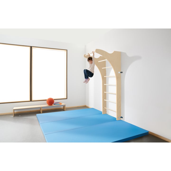 In einem hellen Raum mit großen Fenstern, blauen Turnmatten auf dem Boden und einer niedrigen Bank mit einem orangefarbenen Ball und Ring hängt ein Kind an hölzernen Kletterstangen, die an der Wand befestigt sind.