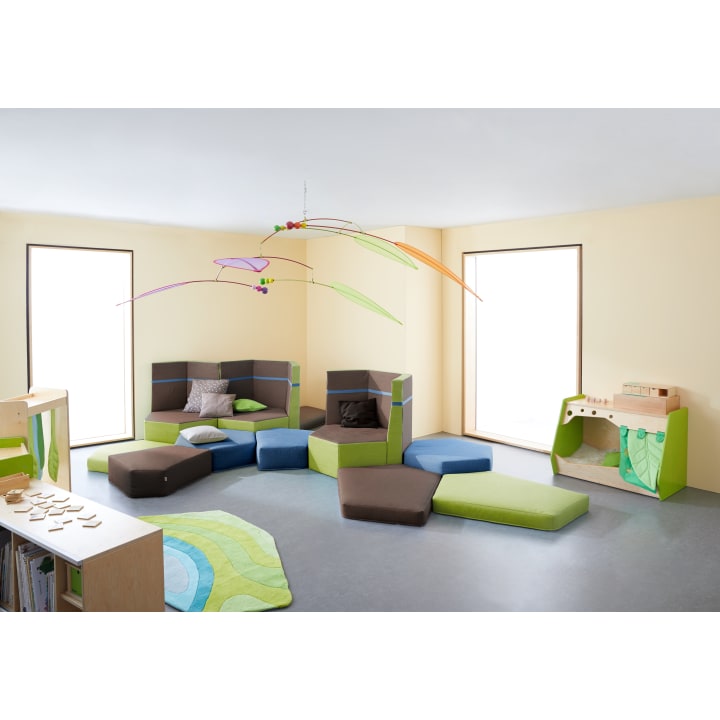 Ein helles Spielzimmer mit großen Fenstern, modularen Schaumstoffsitzmöbeln, einem Teppich, einem hängenden Mobile und Spielmöbeln wie einer Spielzeugküche und Regalen mit Holzspielzeug, alles in Grün-, Braun- und Blautönen.