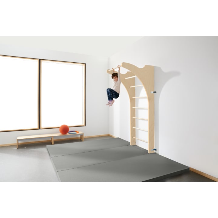In einem hellen Raum mit großen Fenstern, einer Bank, einem orangefarbenen Ball und grauen gepolsterten Matten auf dem Boden hängt ein Kind an einer horizontalen Holzstange, die an einem an der Wand befestigten Klettergerüst angebracht ist.