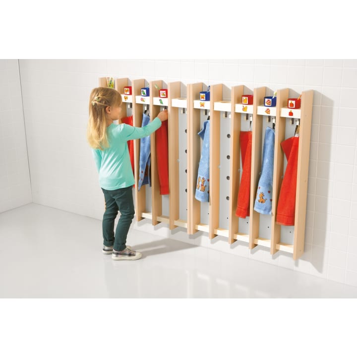 Ein kleines Kind steht vor einer Reihe von Holzfächern mit Haken und hängt ein Handtuch auf. Mehrere Handtücher in Blau und Rot hängen bereits. Jedes Fach hat ein Symbol und ein Regal über dem Haken.