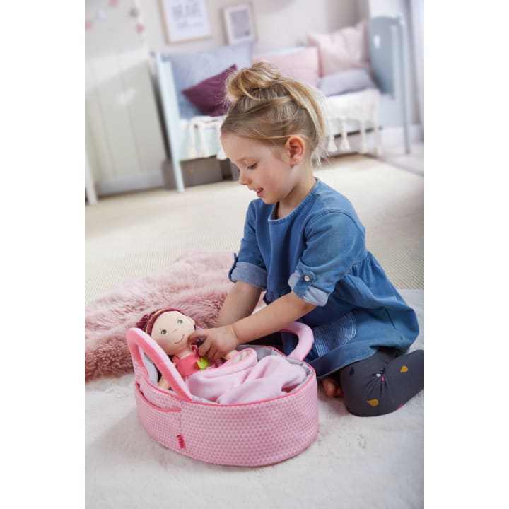 Ein junges Mädchen in einem blauen Kleid sitzt auf einem rosa Teppich und legt eine Puppe in einen rosa Tragekorb. Die Szene spielt in einem gemütlichen, sanft dekorierten Zimmer mit einem Kinderbett und einem Bett im Hintergrund.