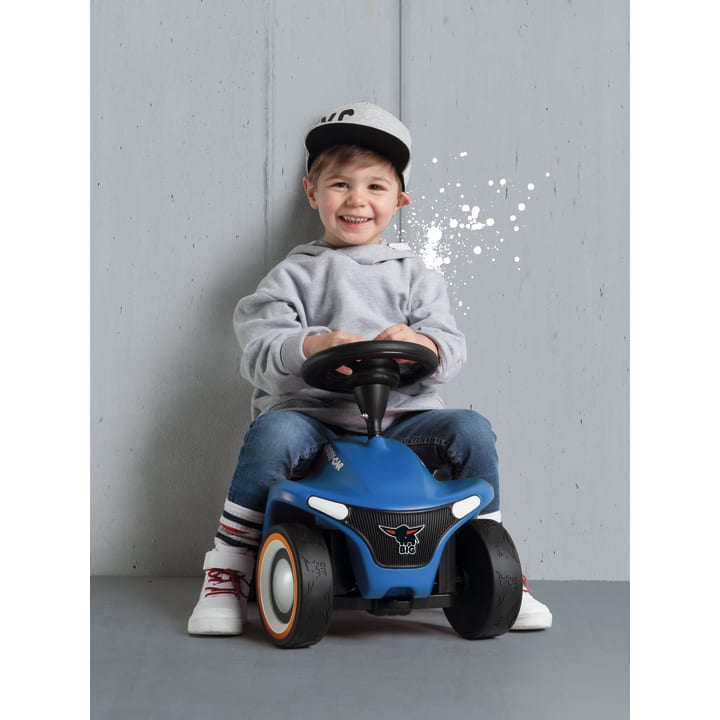 Ein kleines Kind, das einen grauen Kapuzenpullover, blaue Jeans und eine Baseballmütze trägt, sitzt auf einem blauen Spielzeugauto vor einer grauen Wand. Das Kind lächelt und hält das Lenkrad fest.