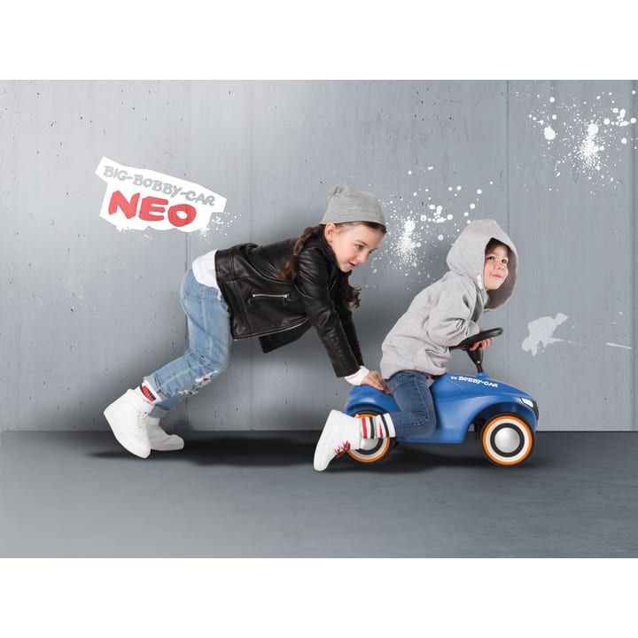 Zwei Kinder spielen in einem Haus vor einer grauen Wand. Ein Kind in einem grauen Kapuzenpulli fährt ein kleines blaues Spielzeugauto, während ein anderes Kind in einer Lederjacke und einer grauen Mütze von hinten schiebt. Die Aufschrift BIG-BOBBY-CAR NEO steht an der Wand.