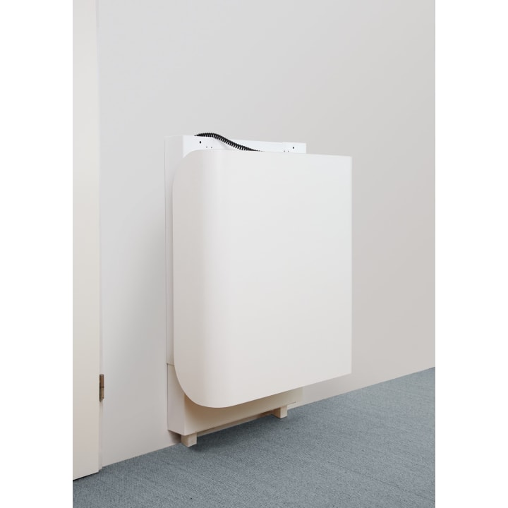 Ein weißer, klappbarer, an der Wand montierter Tisch oder Schreibtisch wird in geschlossener Position vor einer hellen Wand und einem Teppichboden gezeigt. Oben ist ein kleiner Teil eines schwarzen Kabels zu sehen.