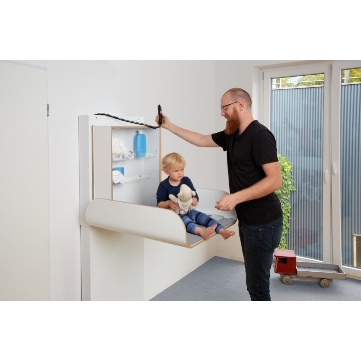 Ein Mann steht neben einem Kleinkind, das auf einem an der Wand befestigten Wickeltisch sitzt. Das Kind hält ein Stofftier in der Hand, und in den Regalen darüber liegen Windeln und Zubehör. Der Mann hält einen schnurgebundenen Telefonhörer in der Hand. Auf dem Boden neben einer Glastür steht ein Spielzeugauto.