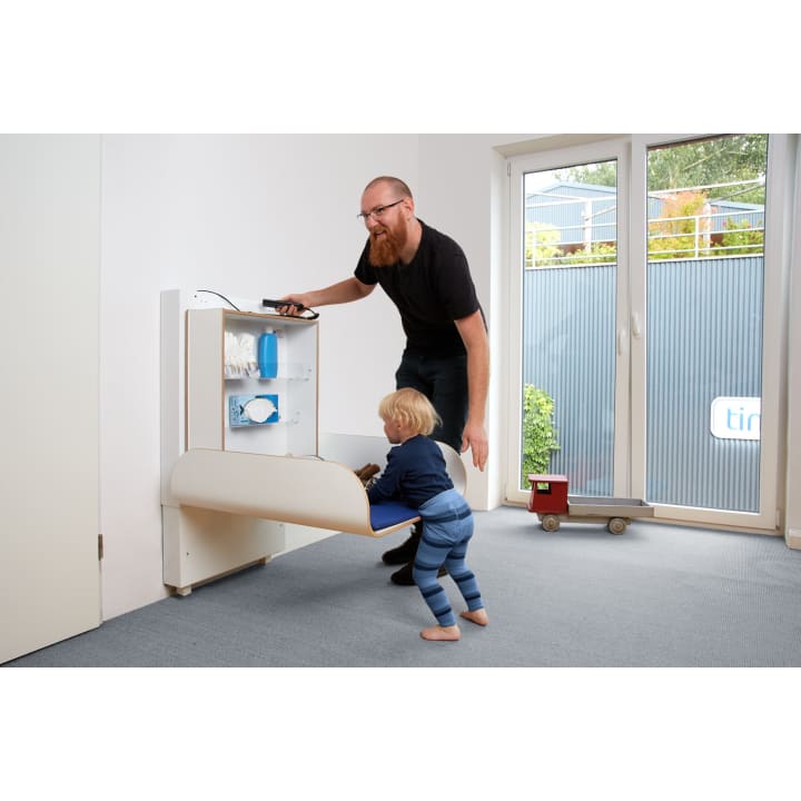 Ein Mann hilft einem Kleinkind in einem Raum mit einer Glastür und einem grauen Teppich, sich neben einem an der Wand montierten Klappwickeltisch aufzustellen. Auf dem Tisch liegen Tücher, Windeln und eine Flasche. Auf dem Boden steht ein kleiner Spielzeuglastwagen.