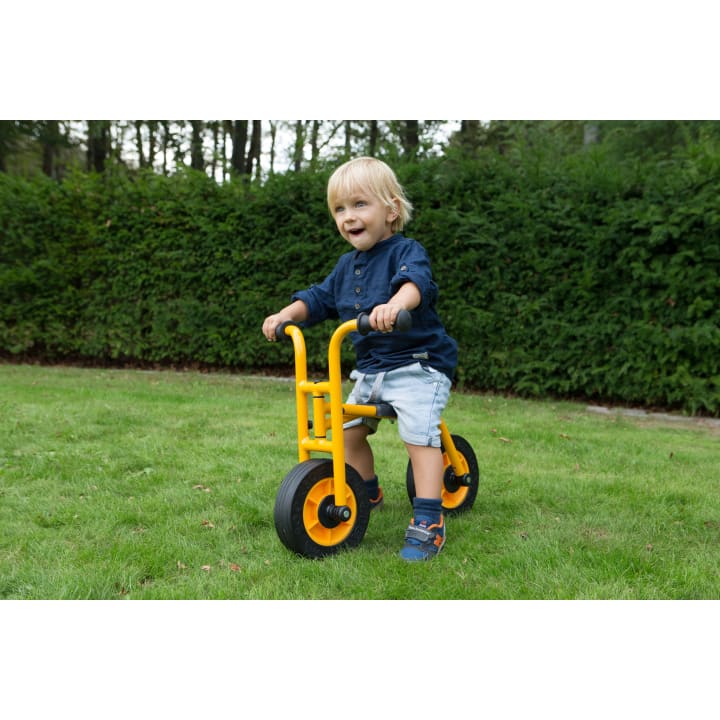 Ein kleines Kind mit blondem Haar, das ein blaues Hemd und helle Shorts trägt, fährt auf einem gelben Dreirad über einen grasbewachsenen Rasen mit dichten grünen Büschen und hohen Bäumen im Hintergrund.