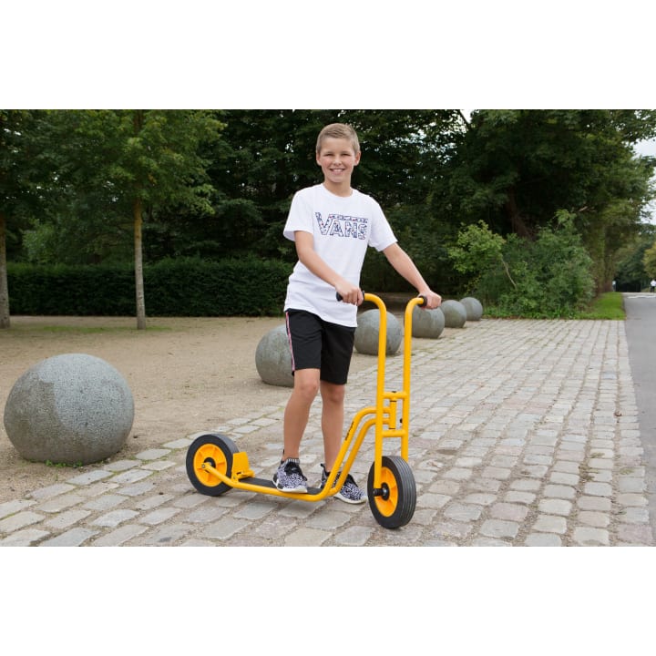 Ein Junge in weißem T-Shirt und schwarzen Shorts steht auf einem gelben Roller auf einem kopfsteingepflasterten Weg, umgeben von Bäumen und großen runden Steinskulpturen.