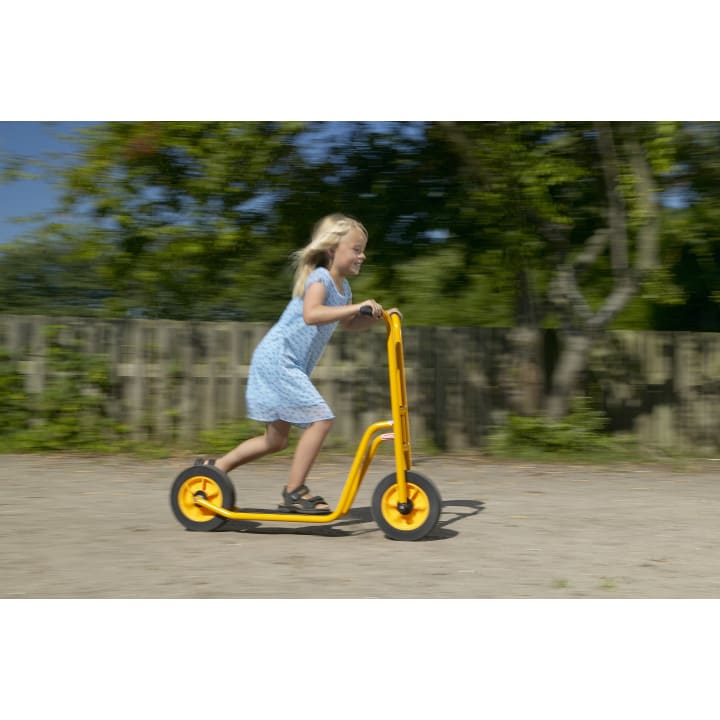Ein junges Mädchen in einem blauen Kleid fährt an einem sonnigen Tag im Freien auf einem gelben Motorroller. Der Hintergrund ist unscharf und suggeriert, dass sie sich schnell bewegt. Hinter ihr sind Bäume und ein Holzzaun zu sehen.