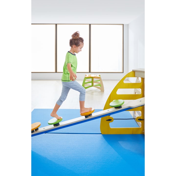 Ein junges Mädchen in einem grünen Hemd und einer hellblauen Hose läuft barfuß auf einem Balance Board in einem hellen Raum mit blauen Matten und großen Fenstern im Hintergrund.