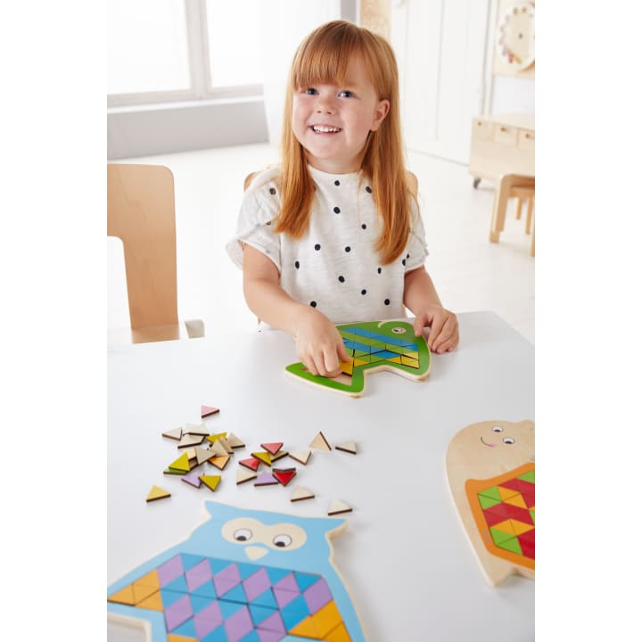 Ein kleines Kind mit roten Haaren und einem weißen gepunkteten Hemd setzt ein hölzernes Eulenpuzzle mit bunten geometrischen Teilen an einem weißen Tisch in einem hellen, kinderfreundlichen Raum zusammen.