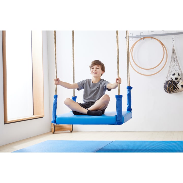 Ein Junge in einem grauen T-Shirt und Shorts sitzt im Schneidersitz auf einer blauen Plattformschaukel in einer Halle und hält sich mit beiden Händen an den Seilen fest. Im Hintergrund sind Gymnastikgeräte und ein Netz mit Bällen zu sehen.