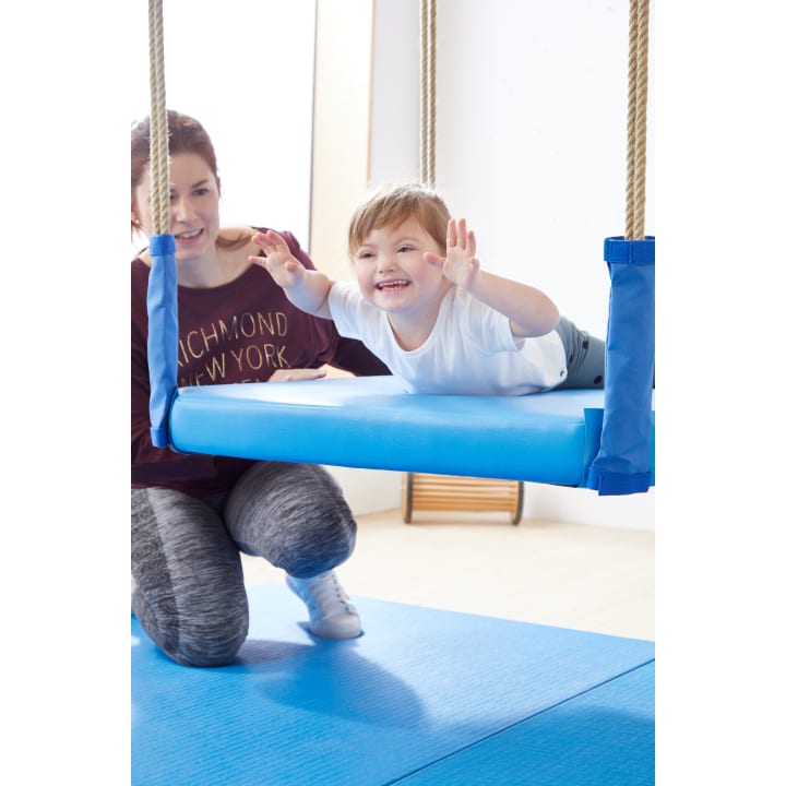 Ein kleines Kind lächelt, während es auf dem Bauch auf einer blauen Schaukel in einer Turnhalle liegt, während ein Erwachsener in der Nähe kniet, um es zu stützen. Beide befinden sich auf blauen Matten in einem hellen Raum.