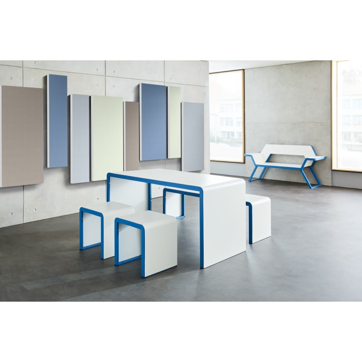 Ein minimalistischer Raum mit einem weißen Tisch und vier passenden Hockern mit blauen Kanten, einer geometrischen weißen Bank und rechteckigen Akustikpaneelen in gedämpften Tönen an der Betonwand. Große Fenster sorgen für natürliches Licht.