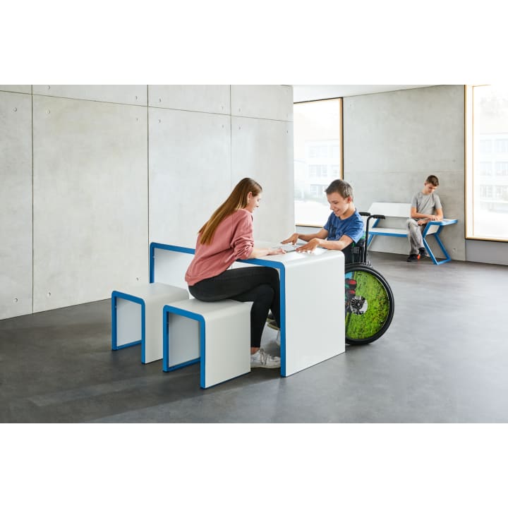 Zwei Personen sitzen an einem weißen Tisch mit blauem Rand; eine sitzt im Rollstuhl. Eine weitere Person arbeitet allein an einem ähnlichen Tisch im Hintergrund. Die Umgebung ist ein modernes, minimalistisches Interieur mit großen Fenstern und Betonwänden.