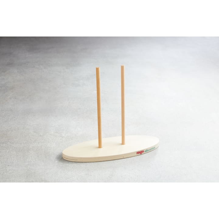 Ein ovaler Holzsockel mit zwei senkrecht stehenden Holzstäben, die auf einer hellgrauen Fläche stehen. Der Sockel hat ein kleines farbiges Logo und einen Text an einer Kante.