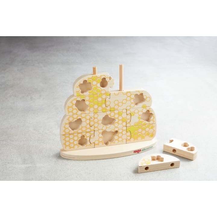 Ein Holzpuzzle in Form eines Bienenstocks mit Bienenwaben und Bienenmotiven. Es fehlen mehrere Teile, zwei Puzzleteile liegen neben dem Puzzle auf dem Boden. Die Marke HABA ist auf dem Sockel sichtbar.