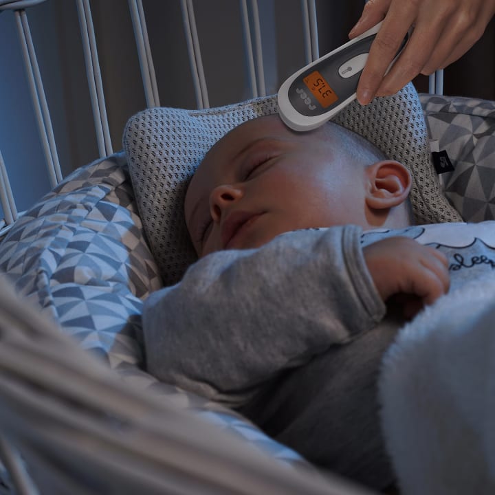 Ein Baby schläft in einem Kinderbett, während jemand ein digitales Thermometer an die Stirn des Babys hält, das einen Messwert anzeigt. Die Bettwäsche für das Kinderbett ist mit einem geometrischen Muster in Grau und Weiß versehen.
