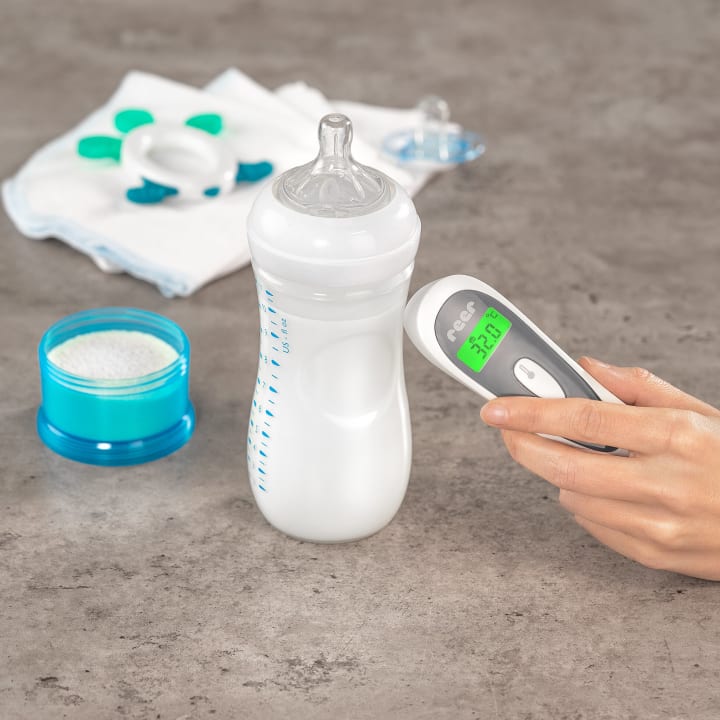 Eine Hand hält ein digitales Thermometer neben einer mit Milch gefüllten Babyflasche. In der Nähe stehen ein Behälter mit Puder, gefaltete Tücher, ein Schnuller und ein Beißring auf einer grauen Arbeitsplatte.