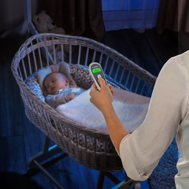 Ein Erwachsener hält in einem schwach beleuchteten Raum ein digitales Infrarot-Thermometer auf ein schlafendes Baby in einem Korbbettchen, das mit einer Decke zugedeckt ist.