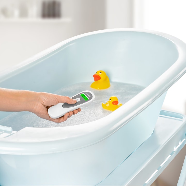Eine Hand hält ein digitales Thermometer über eine mit Wasser gefüllte Babybadewanne. Zwei gelbe Gummi-Enten schwimmen in der Wanne. Die Szene hat einen hellen, sauberen Hintergrund.