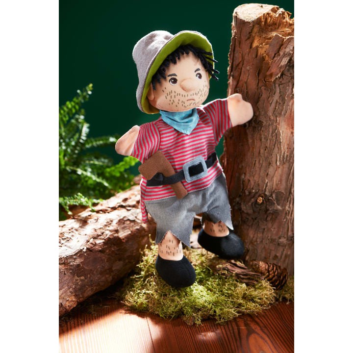 Eine Stoffpuppe, gekleidet als Pirat mit grauem Hut, gestreiftem rotem Hemd, grauen Shorts und schwarzem Gürtel, steht auf moosigem Boden neben Baumstämmen und Grünzeug.
