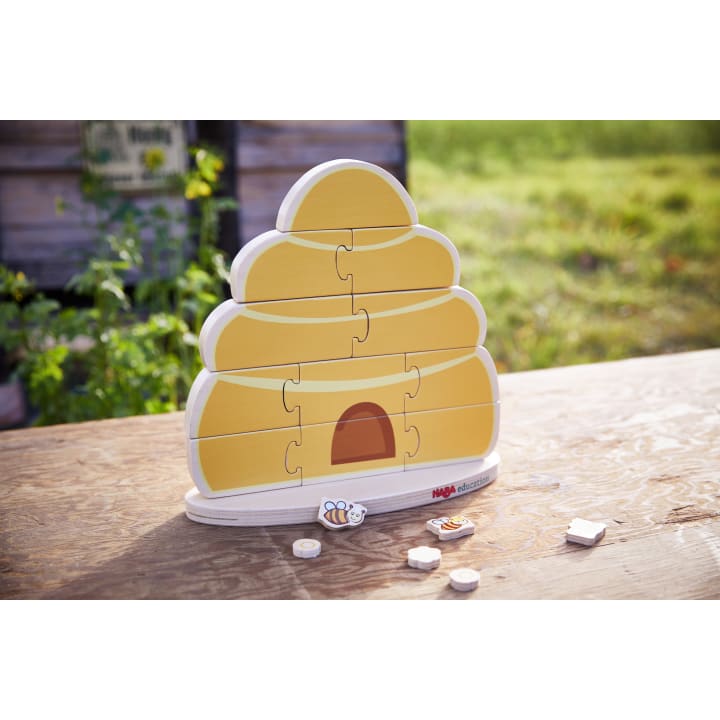 Ein bienenstockförmiges Holzpuzzle mit mehreren fehlenden Teilen liegt auf einem Holztisch im Freien. Einige Puzzleteile, darunter eines mit einer Bienenillustration, sind vor dem Puzzle platziert. Im Hintergrund sind Gras und eine Holzkonstruktion zu sehen.