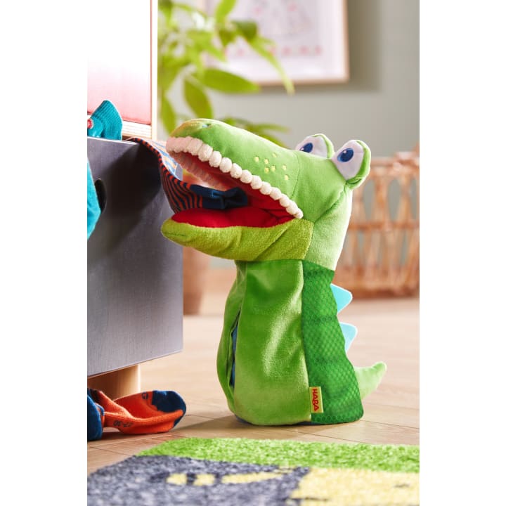 Eine grüne Plüschkrokodilpuppe mit blauen Stacheln hält eine gestreifte Socke im Maul und steht neben einer Schublade, in der weitere Socken zu sehen sind; im Hintergrund befinden sich ein Korb und eine Pflanze.