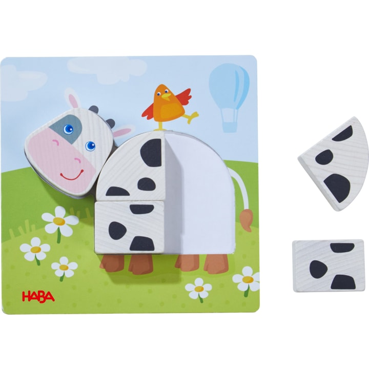 Ein Holzpuzzle mit einer Cartoon-Kuh mit zwei herausnehmbaren schwarz-weißen Teilen und einem lächelnden Gesicht. Ein roter Vogel steht auf dem Rücken der Kuh. Im Hintergrund sind Gras, Blumen und ein blauer Himmel mit einem Heißluftballon zu sehen.
