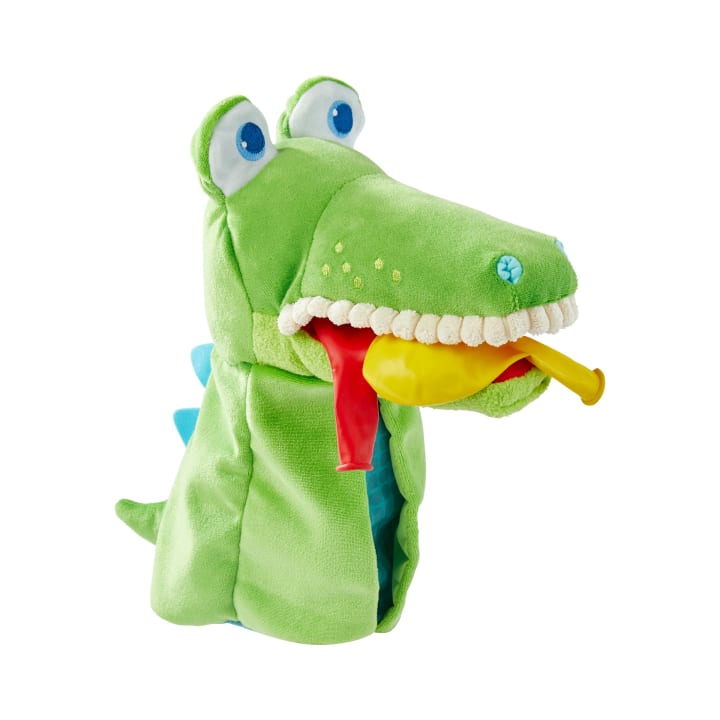 Eine grüne Plüschhandpuppe in Form eines Alligators mit blauen Augen, einer Reihe von Stoffzähnen und einem gelben und roten Luftballon im Maul, dargestellt vor einem weißen Hintergrund.