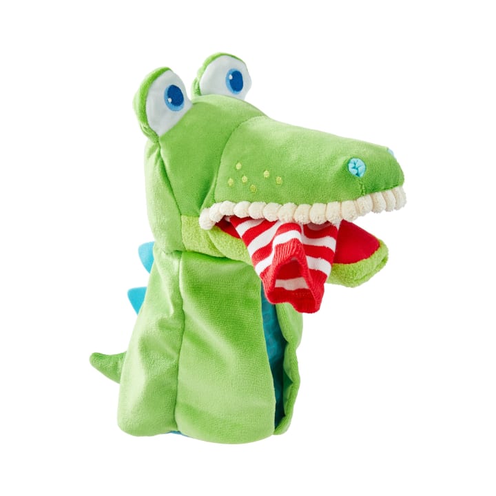 Eine grüne Plüsch-Alligatorpuppe mit blauen Augen und blauen Stacheln hält eine rot-weiß gestreifte Socke im Maul, dargestellt vor einem weißen Hintergrund.