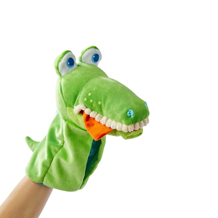 Eine Hand trägt eine grüne Krokodilpuppe aus Plüsch mit weißen Filzzähnen, blau gestickten Nasenlöchern und großen Kulleraugen. Der Hintergrund ist einfarbig weiß.