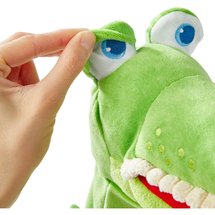Eine Hand hebt das Augenlid eines grünen Plüschtiers mit blauen und weißen Augen, weißen Zähnen und einem roten Mund.