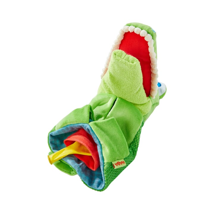 Ein grünes Plüschtier in Form eines Alligators mit offenem Maul, roter Innenseite, weißen Zähnen und einem gelben Beißring an einem Ende, dargestellt auf weißem Hintergrund.