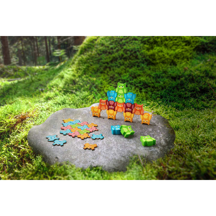 Bunte eulenförmige Spielzeugteile sind auf einem flachen Felsen gestapelt, der von grünem Moos in einem Wald umgeben ist. Weitere Teile, darunter Puzzleformen und Eulen, sind auf dem Felsen verstreut.