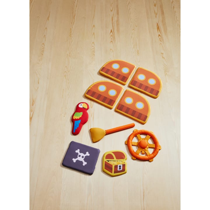 Ein Set aus weichem Plüschspielzeug mit Piratenthematik ist auf einem hellen Holzboden angeordnet. Dazu gehören ein Papagei, eine Schatztruhe, ein Schiffsrad, ein Fernrohr, eine Piratenflagge und vier Teile, die an ein Schiff erinnern.