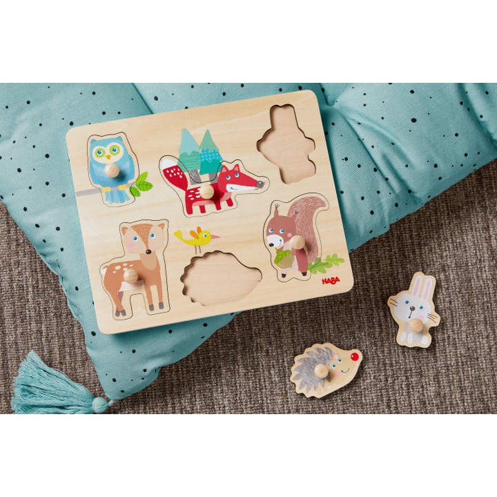 Ein hölzernes Tierpuzzle mit geformten Teilen einer Eule, eines Fuchses, eines Rehs, eines Eichhörnchens, eines Igels und eines Kaninchens liegt auf einem hellblauen Kissen auf einem Teppichboden. Zwei Teile sind herausgenommen und liegen neben dem Puzzlebrett.