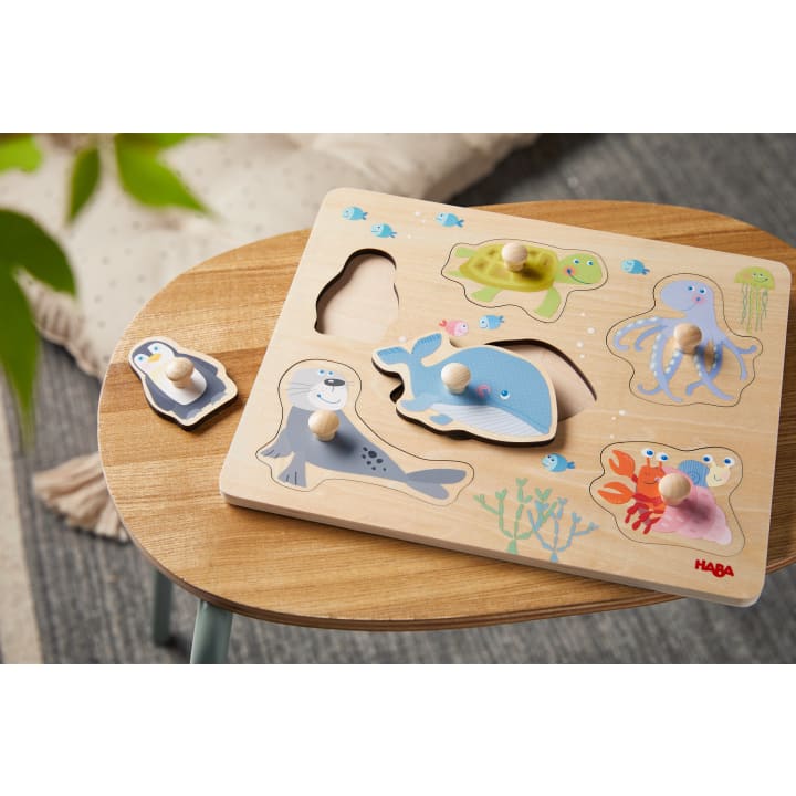 Auf einem kleinen runden Holztisch liegt ein Holzpuzzle mit Meerestieren, darunter ein Pinguin, ein Wal, eine Robbe, eine Schildkröte, ein Oktopus und eine Krabbe. Ein Teil, der Pinguin, ist herausgenommen und liegt neben dem Brett.