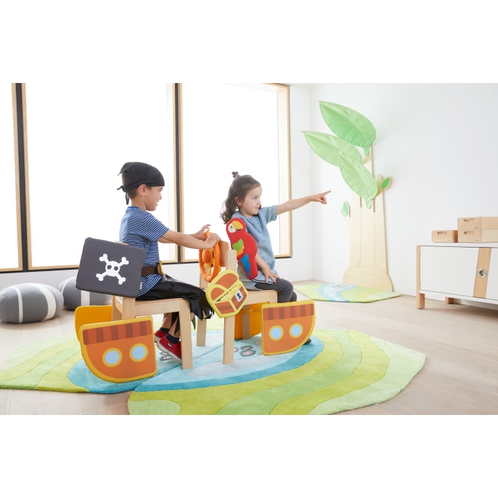 Zwei Kinder sitzen auf als Piratenschiffe dekorierten Stühlen in einem hell erleuchteten Spielzimmer. Ein Kind trägt einen schwarzen Piratenhut, das andere zeigt nach vorne und hält einen Papagei aus Stoff. Es gibt grüne Teppiche und Wanddekorationen in Form von Bäumen.
