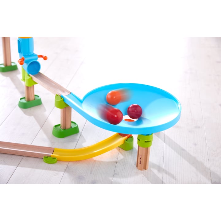 Eine Nahaufnahme einer Kugelbahn auf einem hellen Holzboden. Drei bunte Murmeln drehen sich in einem blauen Trichter, unter dem und um den Trichter herum sind Schienen aus Holz und Kunststoff angebracht.