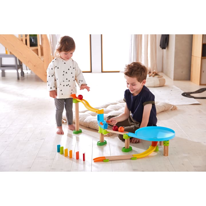 Zwei kleine Kinder spielen mit einer bunten Kugelbahn auf einem hellen Holzboden. Die Bahn verfügt über Rampen, eine Spirale und Dominosteine, die am Ende aufgestellt sind. Im Hintergrund sind Kissen und Holzmöbel zu sehen.
