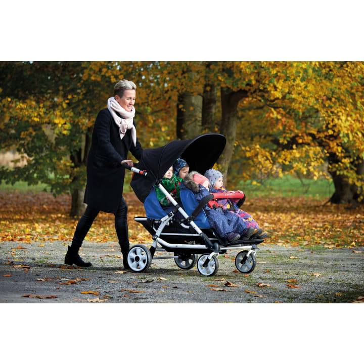 Eine Frau mit dunklem Mantel und Schal schiebt einen großen Kinderwagen mit drei kleinen Kindern, die nebeneinander sitzen. Im Hintergrund sind Bäume mit Herbstlaub und ein mit Laub bedeckter Boden zu sehen.