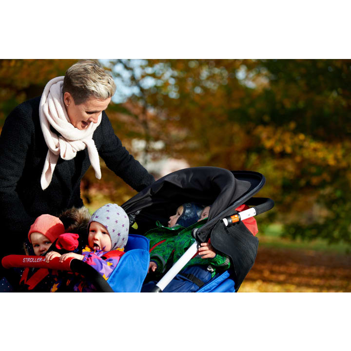 Ein Erwachsener steht im Freien neben einem Kinderwagen mit vier kleinen Kindern, die alle in warme Kleidung gekleidet sind. Der Hintergrund zeigt Herbstlaub mit orangefarbenen und gelben Blättern.